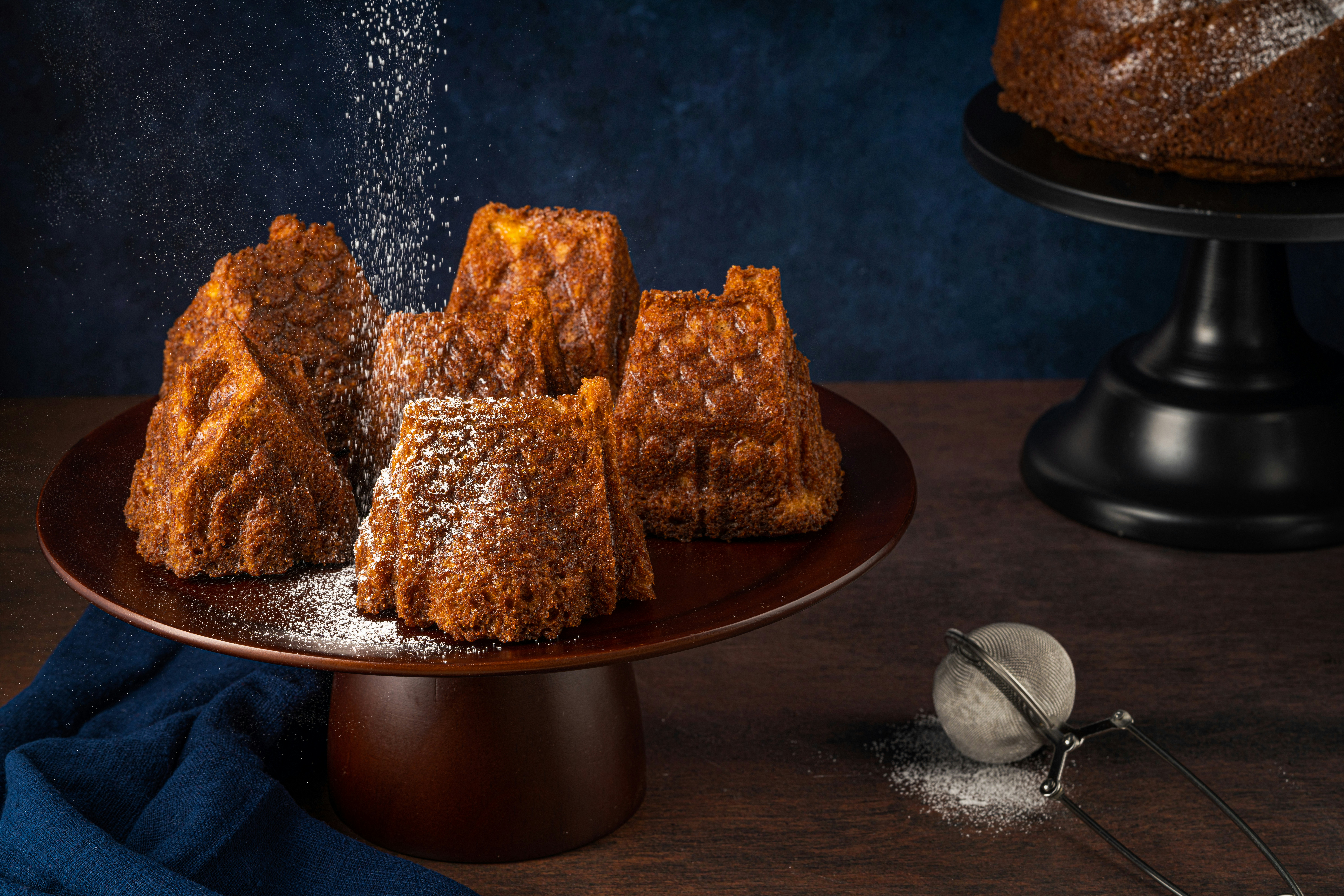 Several intricately shaped brown cakes are arranged on a dark wooden cake stand. A dusting of powdered sugar falls gently over them, adding a light coating to their textured surfaces. A dark blue cloth is draped beside the stand and a metal sieve with some leftover sugar sits on the wooden tabletop. Another cake on a black stand is partially visible in the background, set against a deep blue backdrop.