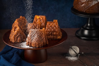 Several intricately shaped brown cakes are arranged on a dark wooden cake stand. A dusting of powdered sugar falls gently over them, adding a light coating to their textured surfaces. A dark blue cloth is draped beside the stand and a metal sieve with some leftover sugar sits on the wooden tabletop. Another cake on a black stand is partially visible in the background, set against a deep blue backdrop.