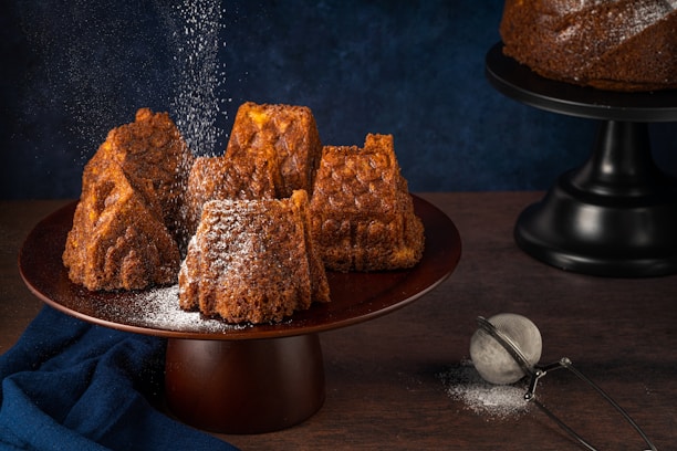 Several intricately shaped brown cakes are arranged on a dark wooden cake stand. A dusting of powdered sugar falls gently over them, adding a light coating to their textured surfaces. A dark blue cloth is draped beside the stand and a metal sieve with some leftover sugar sits on the wooden tabletop. Another cake on a black stand is partially visible in the background, set against a deep blue backdrop.