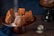 Several intricately shaped brown cakes are arranged on a dark wooden cake stand. A dusting of powdered sugar falls gently over them, adding a light coating to their textured surfaces. A dark blue cloth is draped beside the stand and a metal sieve with some leftover sugar sits on the wooden tabletop. Another cake on a black stand is partially visible in the background, set against a deep blue backdrop.