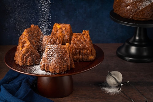 Several intricately shaped brown cakes are arranged on a dark wooden cake stand. A dusting of powdered sugar falls gently over them, adding a light coating to their textured surfaces. A dark blue cloth is draped beside the stand and a metal sieve with some leftover sugar sits on the wooden tabletop. Another cake on a black stand is partially visible in the background, set against a deep blue backdrop.