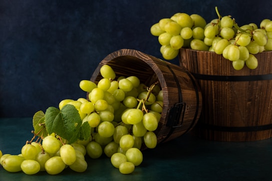 A rustic setup featuring two wooden barrels, one tipped over with a bundle of fresh green grapes spilling out, while another is filled with grapes. The surface is a dark green with a dark blue background, and some green leaves are present among the grapes.