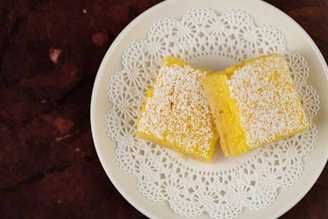 Two yellow square desserts are placed on a white plate with a decorative lace-like pattern. The desserts are topped with a dusting of powdered sugar, and the plate sits on a dark brown textured surface.