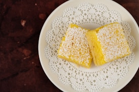 Two yellow square desserts are placed on a white plate with a decorative lace-like pattern. The desserts are topped with a dusting of powdered sugar, and the plate sits on a dark brown textured surface.