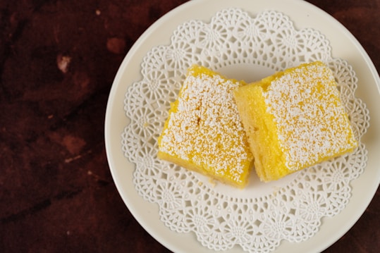 Two yellow square desserts are placed on a white plate with a decorative lace-like pattern. The desserts are topped with a dusting of powdered sugar, and the plate sits on a dark brown textured surface.