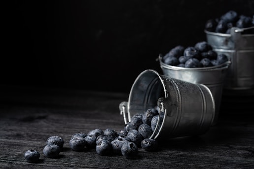 A dark and moody scene with blueberries spilling out of a metal bucket, which is tipped over on a rustic wooden surface. Two additional buckets full of blueberries are visible in the background.