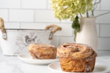 Two freshly baked, golden-brown pastries sit on white plates on a marble surface. In the background, a decorative white container with a bird design and a potted plant with green leaves create a cozy, rustic atmosphere.