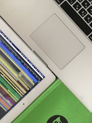 A top-down view of a laptop and a tablet placed diagonally beside each other. The laptop's keyboard and trackpad are visible, while the tablet displays a colorful timeline or graph. A green notebook with a partial logo can also be seen in the bottom right corner.