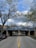 A street view featuring a tree-lined road with cars parked on either side and a blue painted bridge with the words 'TOUHY AVENUE LAKESIDE LIVING' inscribed on it. The sky is partly cloudy, and the surrounding trees display early spring foliage.