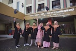 A group of six people dressed in graduation caps and gowns are joyfully throwing their mortarboards into the air. They are standing in front of a building with a sign that indicates it is the Faculty of Da'wa and Communication.