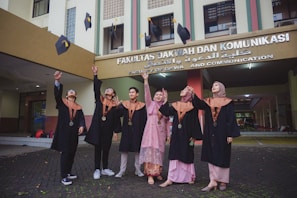 A group of six people dressed in graduation caps and gowns are joyfully throwing their mortarboards into the air. They are standing in front of a building with a sign that indicates it is the Faculty of Da'wa and Communication.