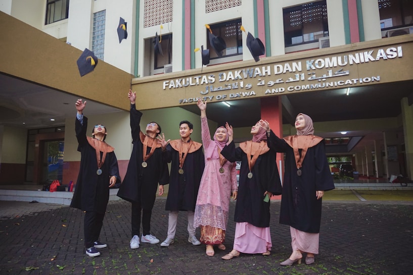 A group of six people dressed in graduation caps and gowns are joyfully throwing their mortarboards into the air. They are standing in front of a building with a sign that indicates it is the Faculty of Da'wa and Communication.