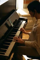 A person is playing the piano with focused attention, framed in soft lighting. The scene appears to be indoors with sunlight streaming through a window, creating a peaceful and intimate atmosphere.