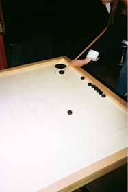 A person is playing carrom, holding a striker with a cue on a wooden carrom board. Several black carrom pieces are arranged near the corner pocket.