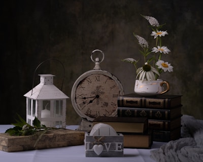 An antique clock with Roman numerals rests against a backdrop of dark tones. Next to it, a stack of vintage books is topped with a teacup filled with white daisies and greenery. A white lantern with a candle is positioned on a decorative box, and a wooden block with the word 'LOVE' sits in the foreground alongside a few scattered leaves and a textured fabric.