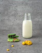 A glass bottle filled with milk stands upright on a concrete-textured surface. To the left, a small stack of green cookies rests, topped with colorful sprinkles. In front, a bitten yellow cookie with sprinkles adds a playful and casual element.