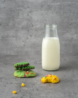 A glass bottle filled with milk stands upright on a concrete-textured surface. To the left, a small stack of green cookies rests, topped with colorful sprinkles. In front, a bitten yellow cookie with sprinkles adds a playful and casual element.