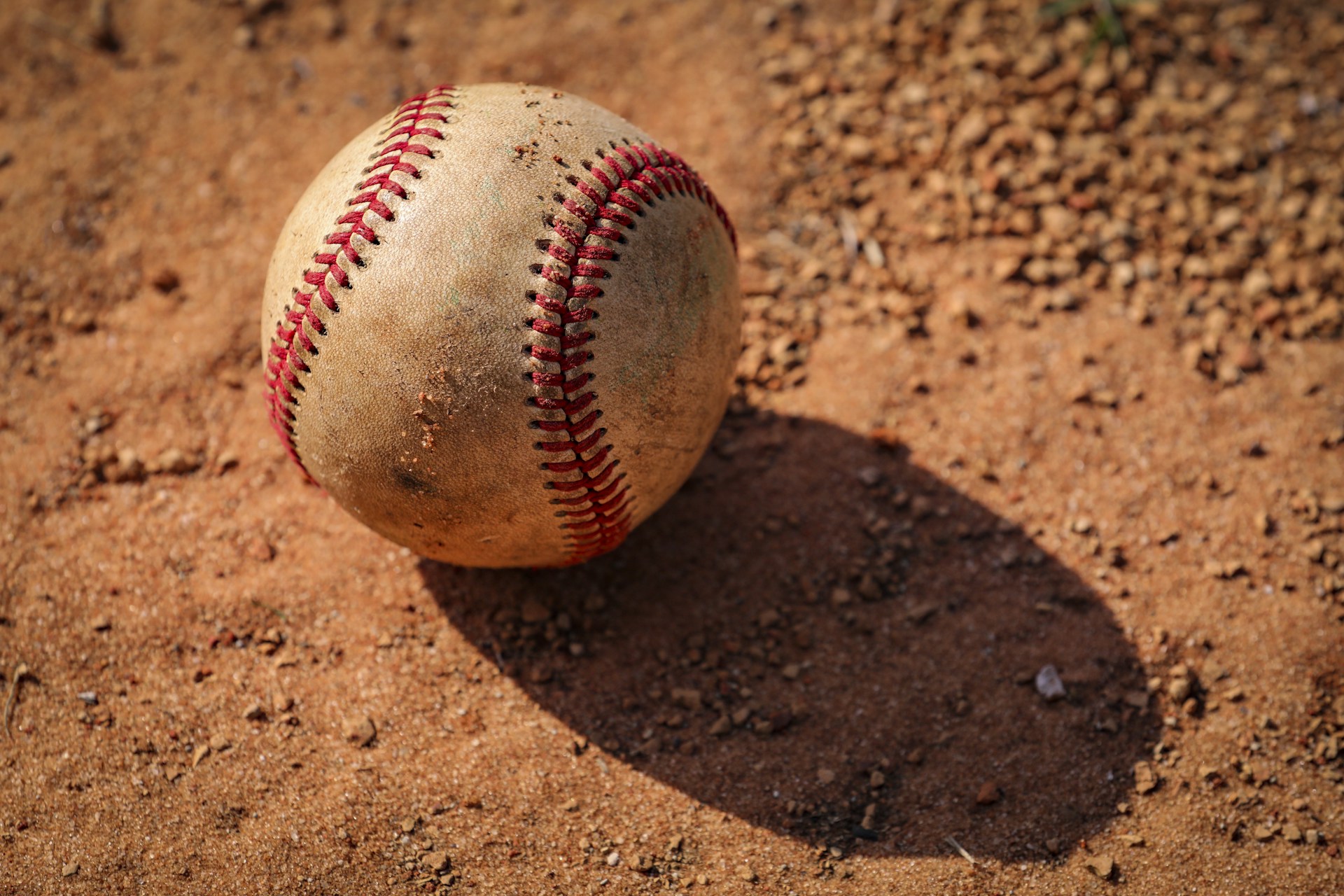 A worn-out baseball rests on a textured dirt surface, casting a shadow to its side. The baseball's red stitching is prominent against its weathered, beige exterior, contrasting with the rough, sandy ground underneath.