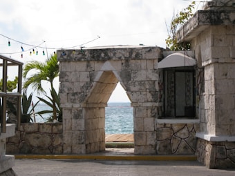 A stone archway frames a view of the ocean beyond. The structure is weathered, suggesting a seaside location. Tropical plants can be seen to the left, and decorative lights hang above the arch. The ocean is visible through the arch, adding a serene touch to the scene.