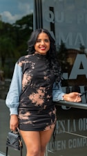A woman with long black hair and a bright smile is standing in front of a glass wall with text on it. She is wearing a stylish black and beige patterned dress with light blue sleeves, holding a black handbag in one hand and sunglasses in the other.