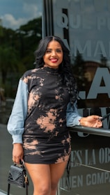 A woman with long black hair and a bright smile is standing in front of a glass wall with text on it. She is wearing a stylish black and beige patterned dress with light blue sleeves, holding a black handbag in one hand and sunglasses in the other.