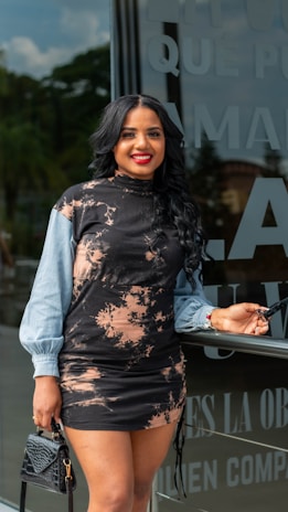 A woman with long black hair and a bright smile is standing in front of a glass wall with text on it. She is wearing a stylish black and beige patterned dress with light blue sleeves, holding a black handbag in one hand and sunglasses in the other.