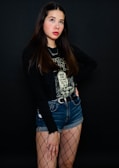 A person with long, dark brown hair stands against a dark background. They are wearing a black graphic T-shirt, a black jacket, denim shorts, and fishnet stockings.