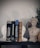 A shelf displays a collection of books on pharmacology, flanked by two white statue bookends in the shape of classical heads and a stone bust of a classical figure. A small green plant in a woven basket adds a touch of natural color.