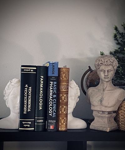 A shelf displays a collection of books on pharmacology, flanked by two white statue bookends in the shape of classical heads and a stone bust of a classical figure. A small green plant in a woven basket adds a touch of natural color.