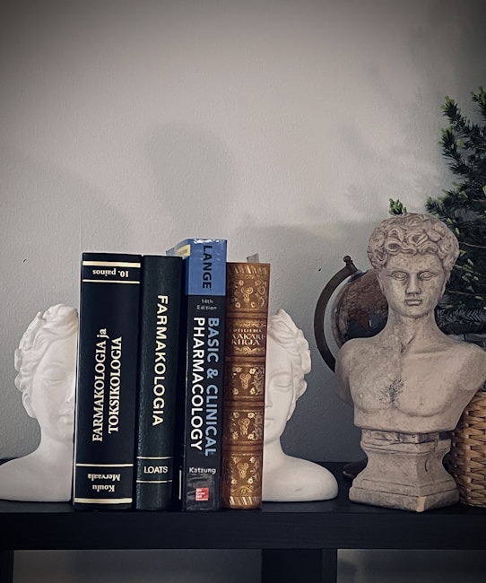 A shelf displays a collection of books on pharmacology, flanked by two white statue bookends in the shape of classical heads and a stone bust of a classical figure. A small green plant in a woven basket adds a touch of natural color.
