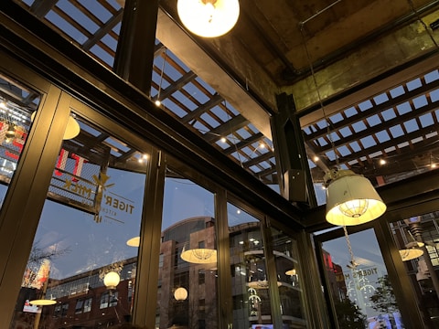 The image captures an interior space with large glass windows, revealing an urban environment outside. The ceiling features an industrial design with exposed beams and hanging light fixtures. Reflected in the glass are various bright lights and signage from outside. The atmosphere suggests a commercial or dining area.