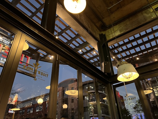 The image captures an interior space with large glass windows, revealing an urban environment outside. The ceiling features an industrial design with exposed beams and hanging light fixtures. Reflected in the glass are various bright lights and signage from outside. The atmosphere suggests a commercial or dining area.