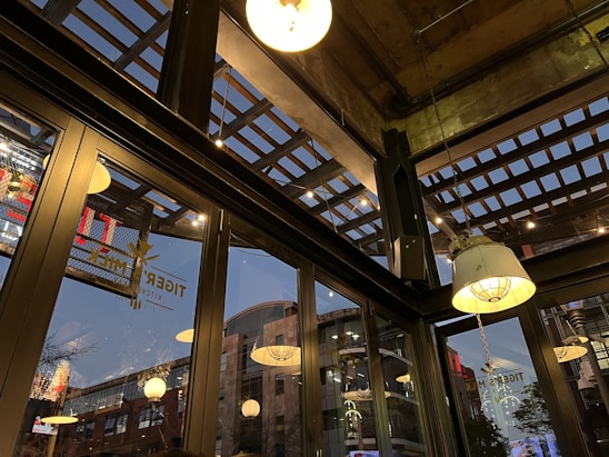 The image captures an interior space with large glass windows, revealing an urban environment outside. The ceiling features an industrial design with exposed beams and hanging light fixtures. Reflected in the glass are various bright lights and signage from outside. The atmosphere suggests a commercial or dining area.