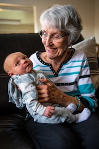 An elderly woman with white hair and glasses is holding a newborn baby in her arms. The woman is smiling warmly, looking at the baby with affection. The baby, wrapped in a soft, patterned blanket, is gazing up with wide eyes. They are seated on a dark sofa in a well-lit room, creating a cozy and intimate atmosphere.