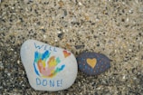 Two painted stones are placed on a textured surface. One stone features colorful abstract designs with the words 'WELL DONE!' written in blue, and a small red heart. The other stone is dark with a yellow heart and small dotted patterns in blue and orange.