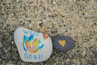 Two painted stones are placed on a textured surface. One stone features colorful abstract designs with the words 'WELL DONE!' written in blue, and a small red heart. The other stone is dark with a yellow heart and small dotted patterns in blue and orange.