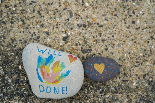 Two painted stones are placed on a textured surface. One stone features colorful abstract designs with the words 'WELL DONE!' written in blue, and a small red heart. The other stone is dark with a yellow heart and small dotted patterns in blue and orange.