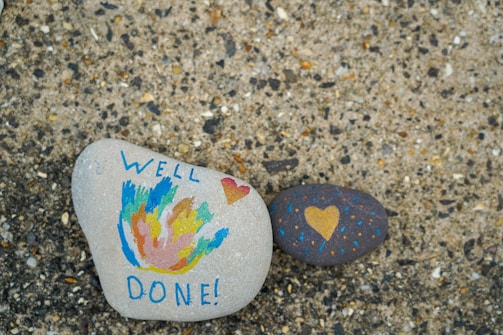Two painted stones are placed on a textured surface. One stone features colorful abstract designs with the words 'WELL DONE!' written in blue, and a small red heart. The other stone is dark with a yellow heart and small dotted patterns in blue and orange.