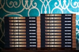 Two stacks of books are arranged, each book with a title that reads 'Team Lead Succeed.' The author's name, Nick Fewings, is visible on the spines. The books are neatly placed on a wooden surface, in front of a decorative background with intricate teal and cream patterns.