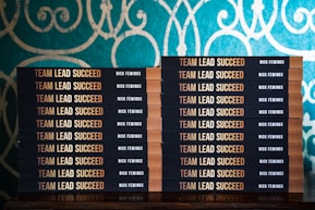 Two stacks of books are arranged, each book with a title that reads 'Team Lead Succeed.' The author's name, Nick Fewings, is visible on the spines. The books are neatly placed on a wooden surface, in front of a decorative background with intricate teal and cream patterns.