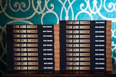 Two stacks of books are arranged, each book with a title that reads 'Team Lead Succeed.' The author's name, Nick Fewings, is visible on the spines. The books are neatly placed on a wooden surface, in front of a decorative background with intricate teal and cream patterns.