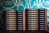 Two stacks of books are arranged, each book with a title that reads 'Team Lead Succeed.' The author's name, Nick Fewings, is visible on the spines. The books are neatly placed on a wooden surface, in front of a decorative background with intricate teal and cream patterns.