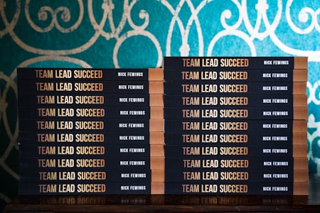 Two stacks of books are arranged, each book with a title that reads 'Team Lead Succeed.' The author's name, Nick Fewings, is visible on the spines. The books are neatly placed on a wooden surface, in front of a decorative background with intricate teal and cream patterns.