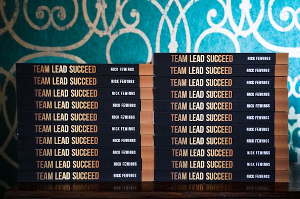 Two stacks of books are arranged, each book with a title that reads 'Team Lead Succeed.' The author's name, Nick Fewings, is visible on the spines. The books are neatly placed on a wooden surface, in front of a decorative background with intricate teal and cream patterns.
