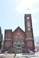 A large brick church with a central clock tower and gothic architecture. The building features multiple tall arched windows and a Canadian flag flying at the top of the tower. There is a sign at the entrance that reads 'Food for the Soul!'. The sky is clear with a few clouds.