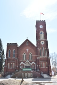 A large brick church with a central clock tower and gothic architecture. The building features multiple tall arched windows and a Canadian flag flying at the top of the tower. There is a sign at the entrance that reads 'Food for the Soul!'. The sky is clear with a few clouds.