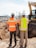 Two construction workers wearing orange safety vests with reflective strips and helmets stand side by side, overlooking an excavation site. One worker is wearing a white helmet and the other is wearing a colorful helmet with stickers. In the background, there is a bulldozer and a fenced-off area with various construction materials. Mountains and a clear sky can be seen in the distance.