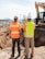 Two construction workers wearing orange safety vests with reflective strips and helmets stand side by side, overlooking an excavation site. One worker is wearing a white helmet and the other is wearing a colorful helmet with stickers. In the background, there is a bulldozer and a fenced-off area with various construction materials. Mountains and a clear sky can be seen in the distance.