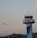 An airplane is taking off into a clear sky, ascending near an airport control tower. The control tower is modern, with a sleek, metallic finish and various antennas on top. In the background, there are silhouettes of trees and distant buildings.