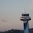 An airplane is taking off into a clear sky, ascending near an airport control tower. The control tower is modern, with a sleek, metallic finish and various antennas on top. In the background, there are silhouettes of trees and distant buildings.